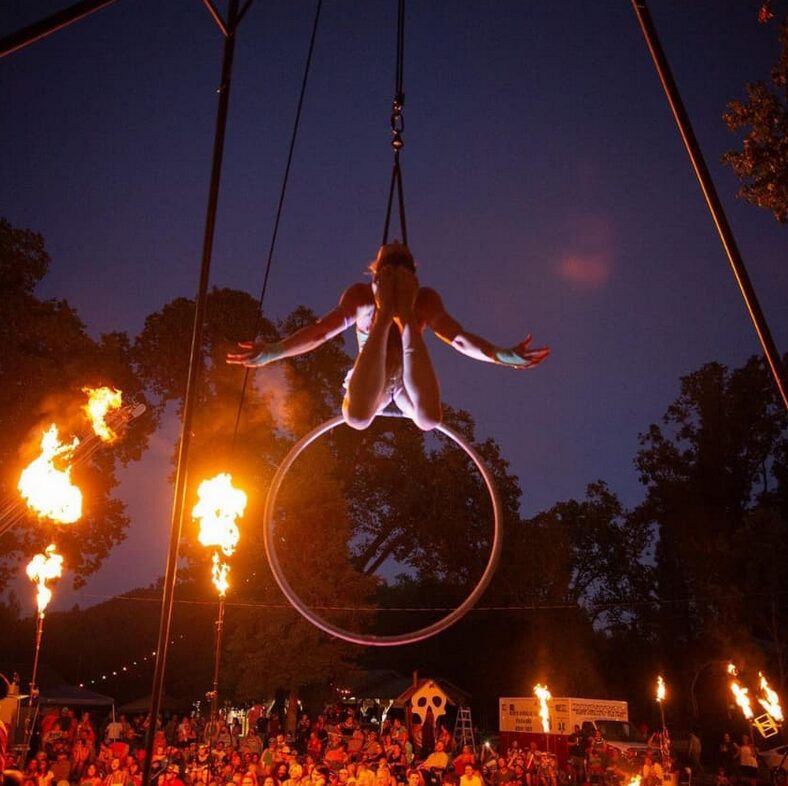 Aerial hoop performer suspended above a fire-lit crowd at dusk